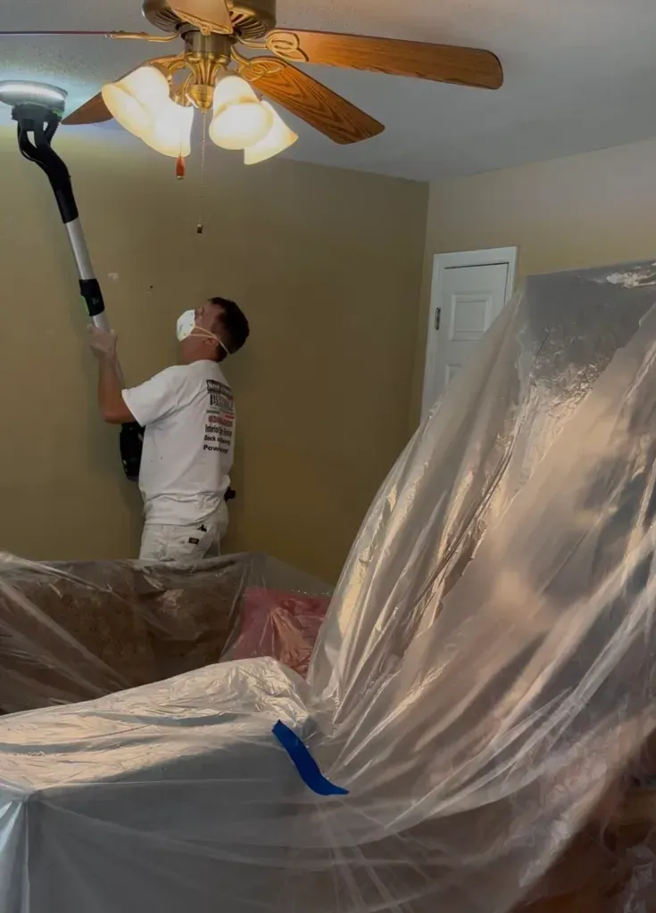Person sanding a ceiling, wearing a mask, with plastic sheeting covering furniture in a room. Person sanding a ceiling, wearing a mask, with plastic sheeting covering furniture in a room.