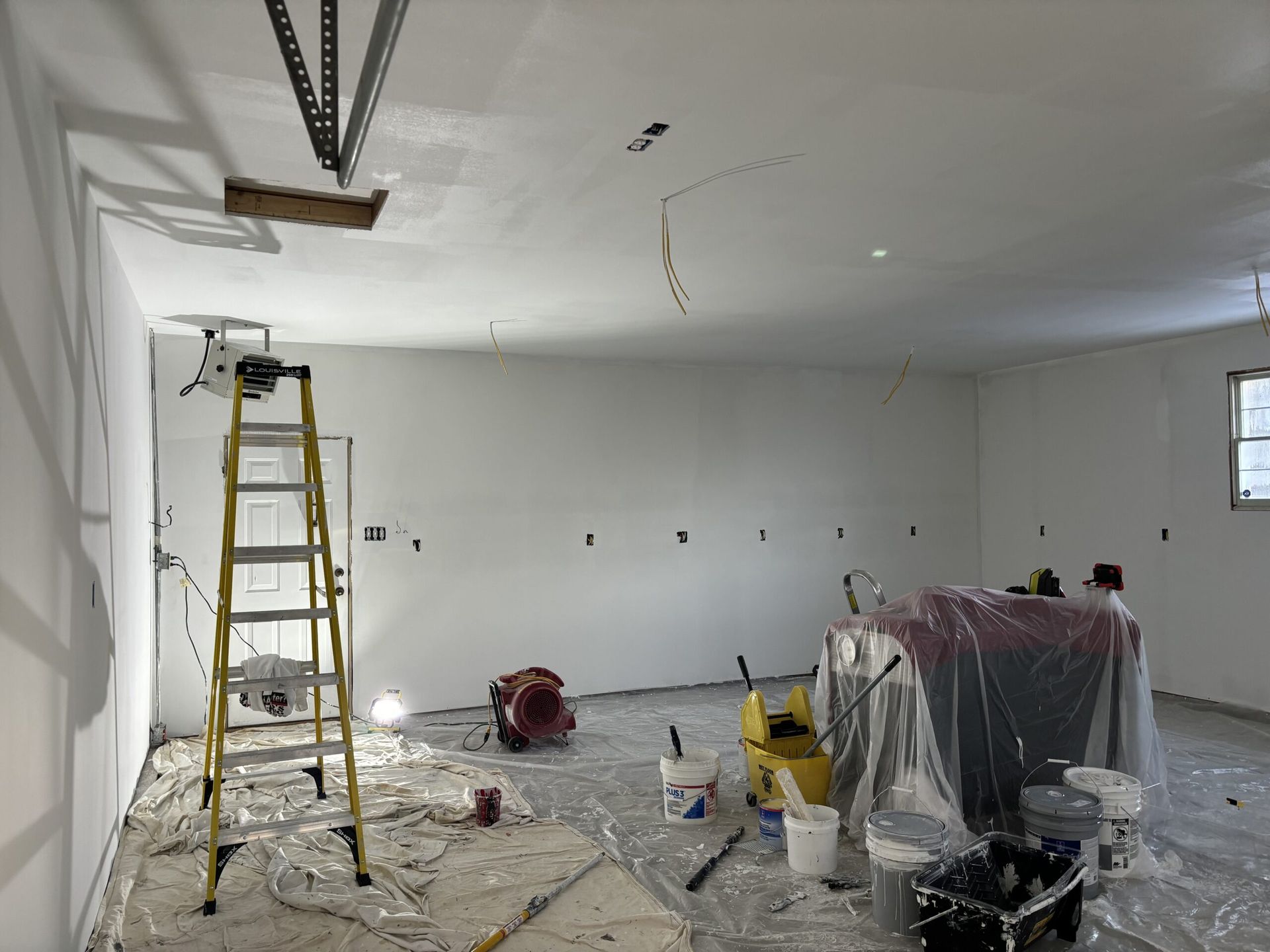 Interior view of a garage being renovated; white walls, ladder, paint supplies, and equipment covered in plastic.