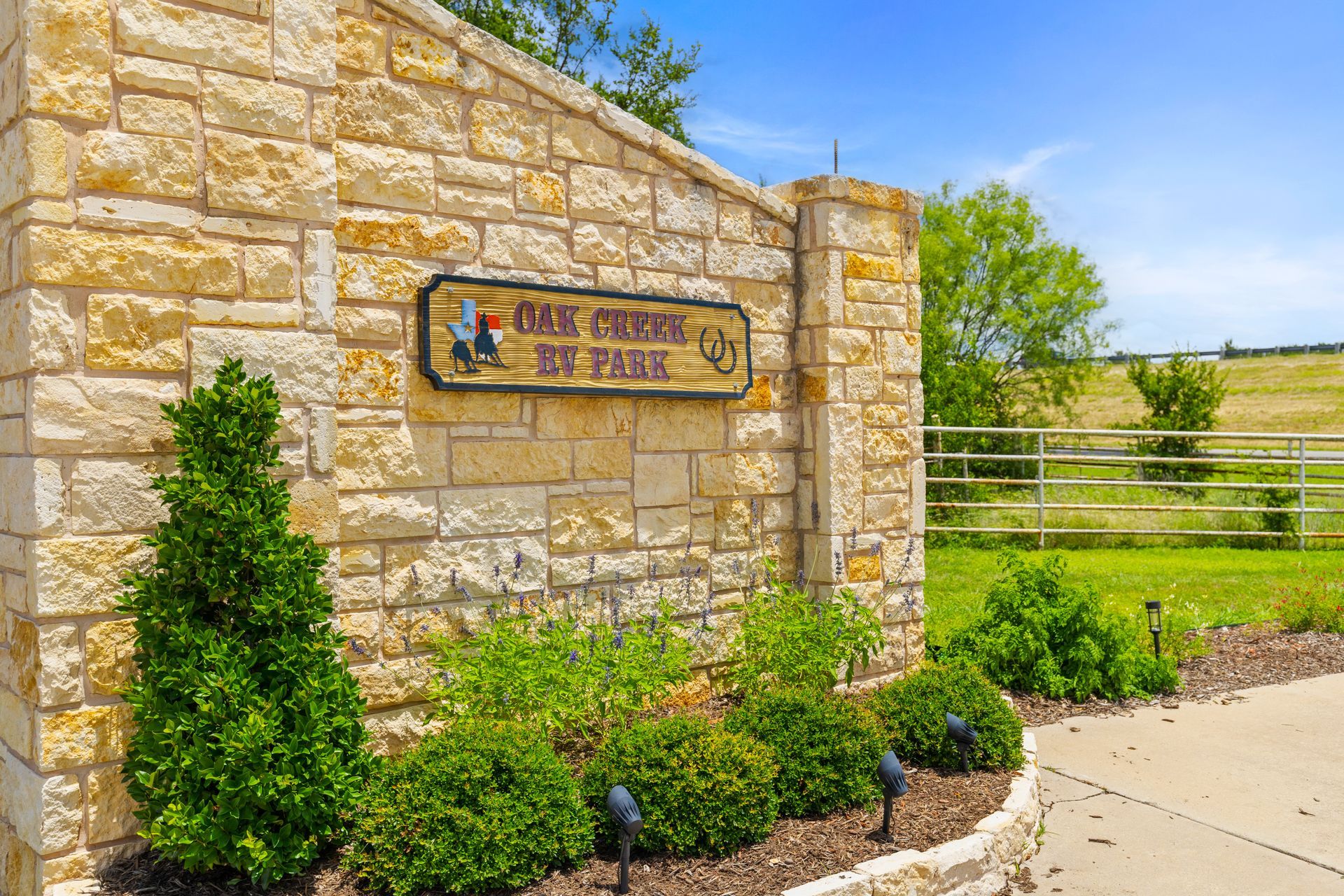 Stone entrance sign for a park beside a sidewalk, with landscaping and a field in the background