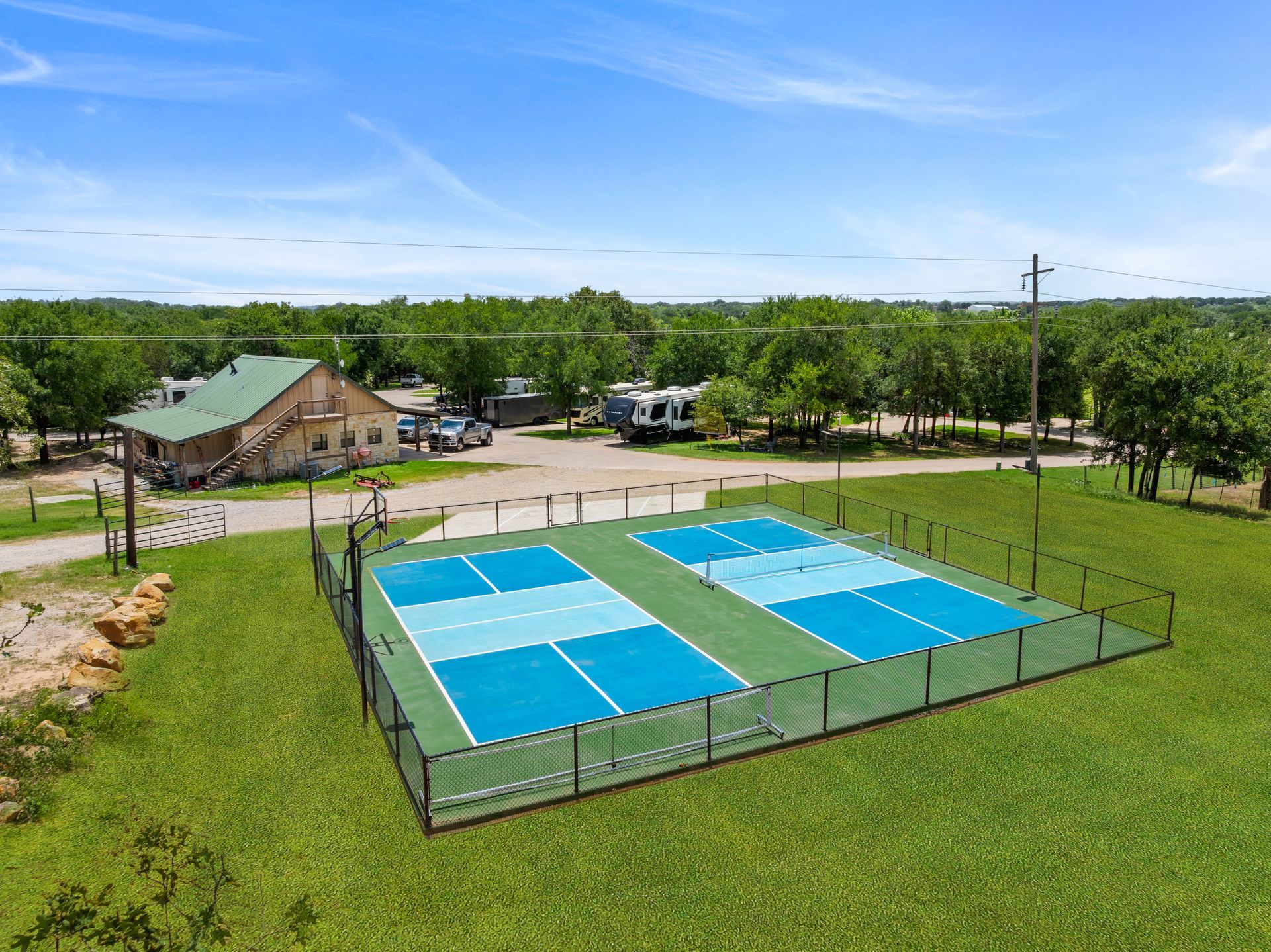 Aerial view of two blue tennis courts beside a grassy field and small building under a clear sky