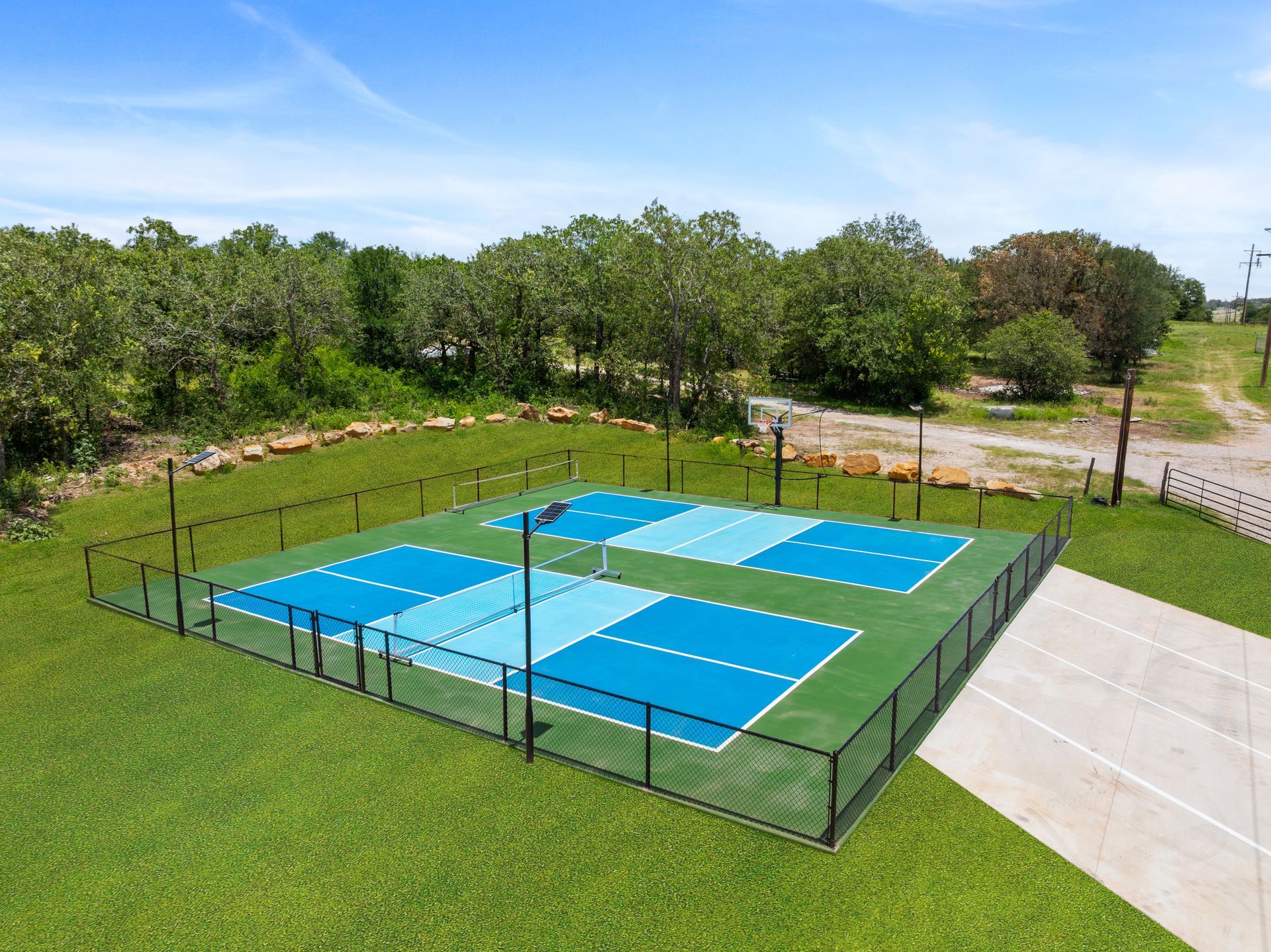 Empty blue outdoor tennis court surrounded by grass, trees, and a paved path