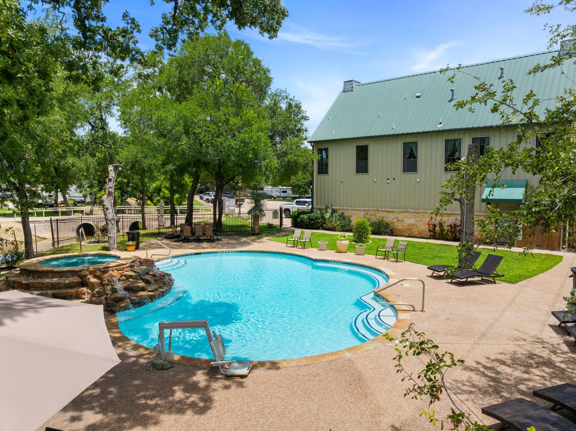 Backyard swimming pool beside a beige house with green roof and trees on a sunny day