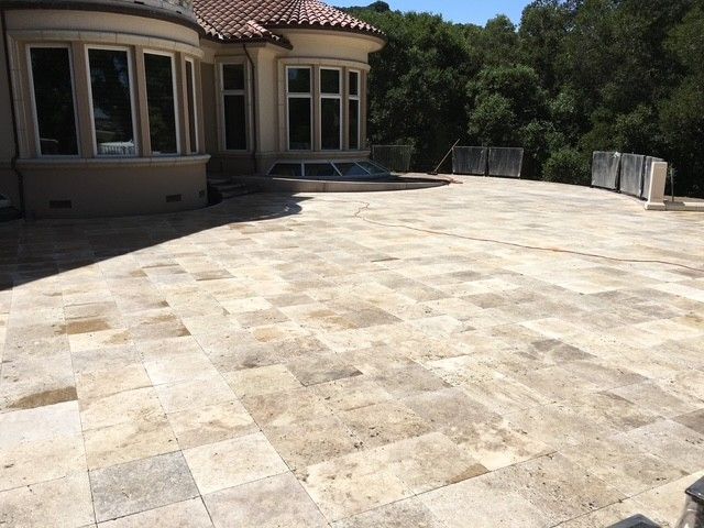 Stone patio with building and trees. Light-colored rectangular pavers cover the ground. Large windows visible.