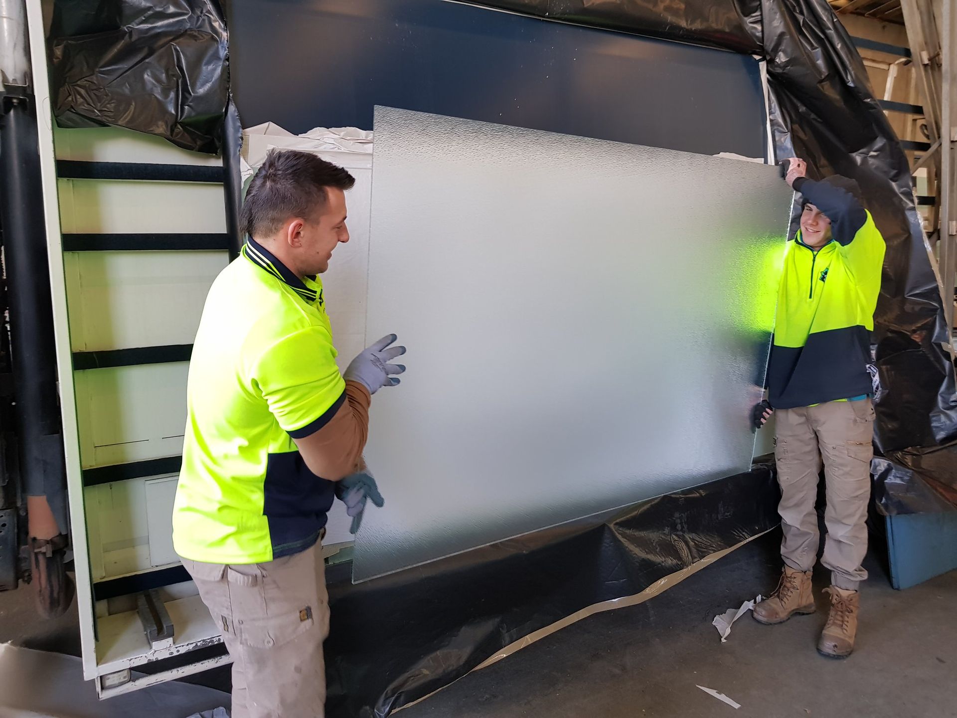 Two workers in neon vests install a large frosted glass panel.