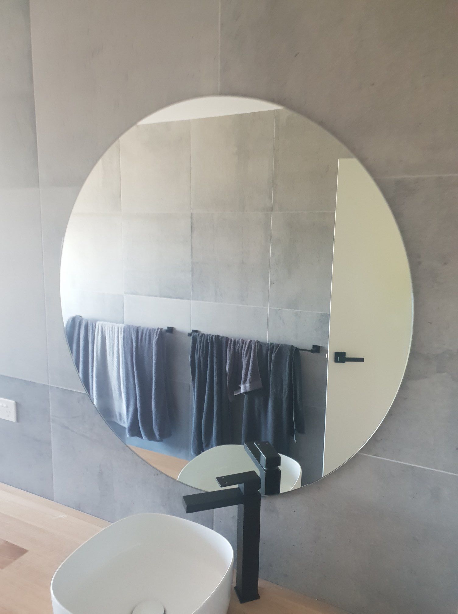 Round mirror over a bathroom sink, reflecting towels, a door, and faucet. Gray tile wall.