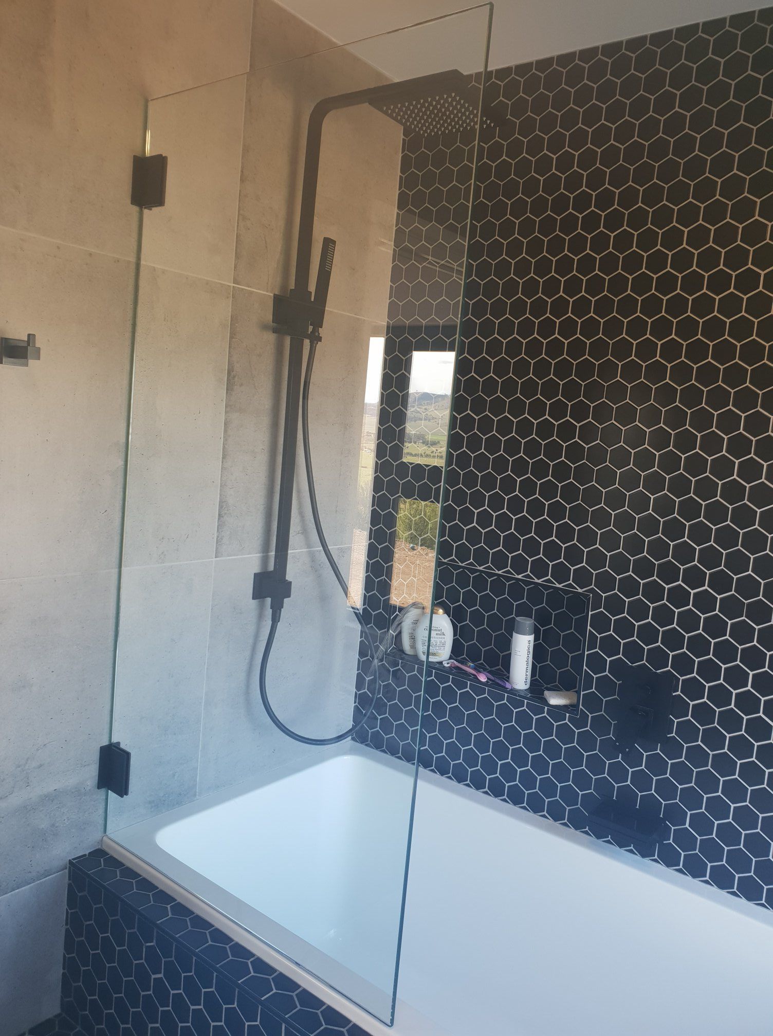 Bathroom with black shower fixtures, glass partition, and black mosaic tile accent wall. White bathtub.