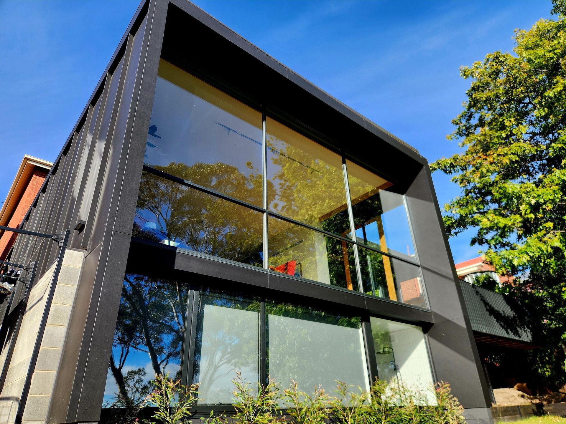 Modern black and glass house with large windows reflecting trees, under a blue sky.