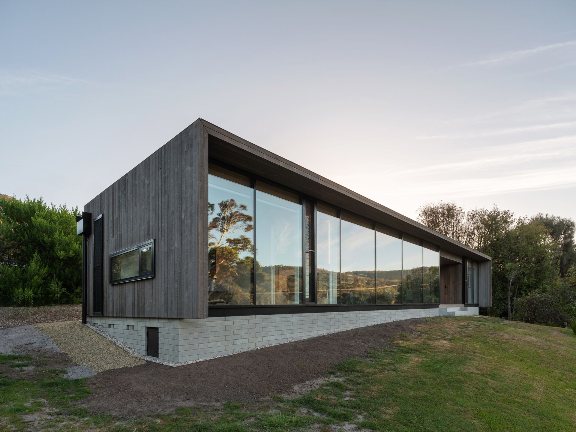 Modern wood-clad house with floor-to-ceiling windows and concrete base, set on a grassy hill.