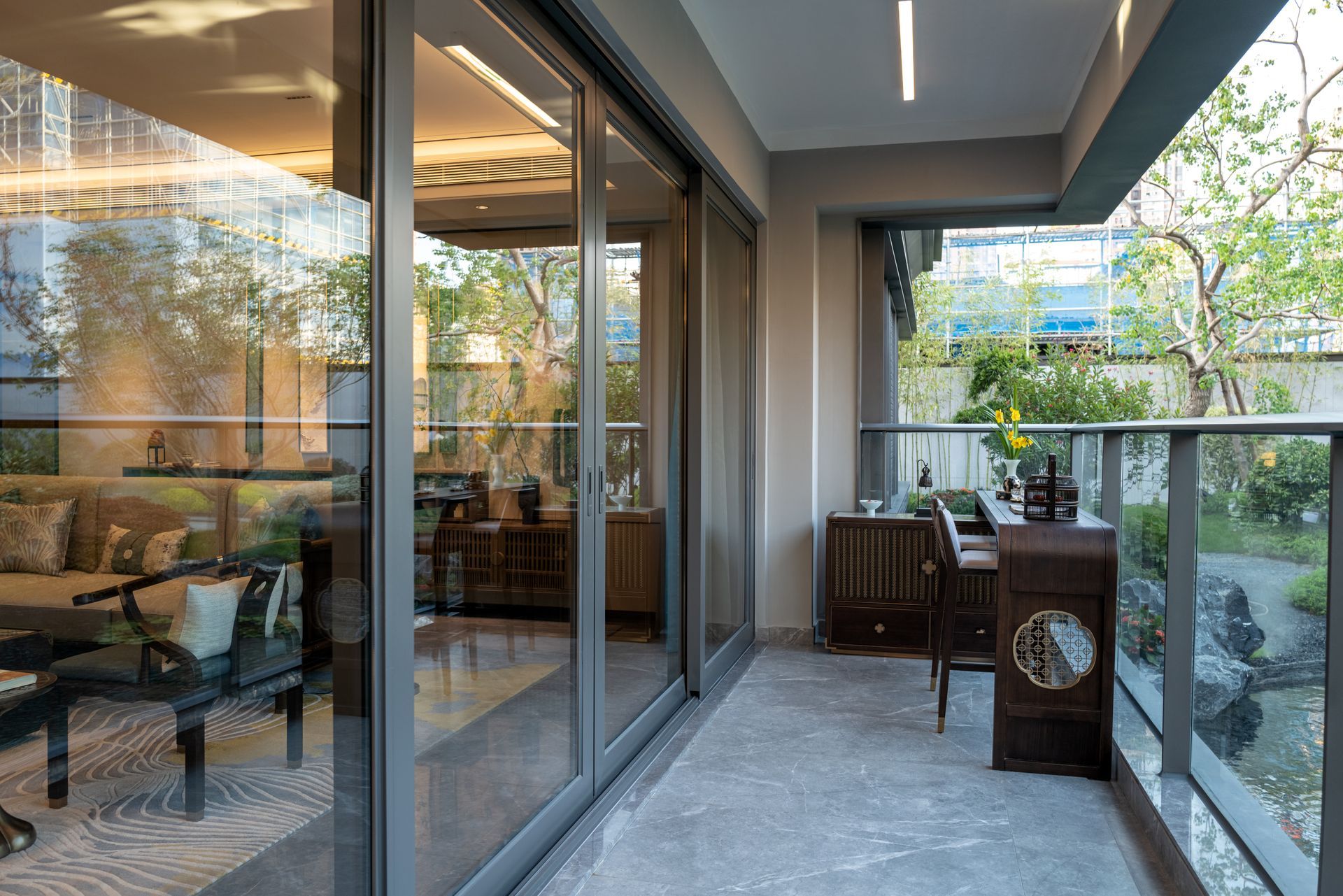 Balcony with glass railing and sliding doors, overlooking a garden. Wooden furniture and contemporary interior visible.