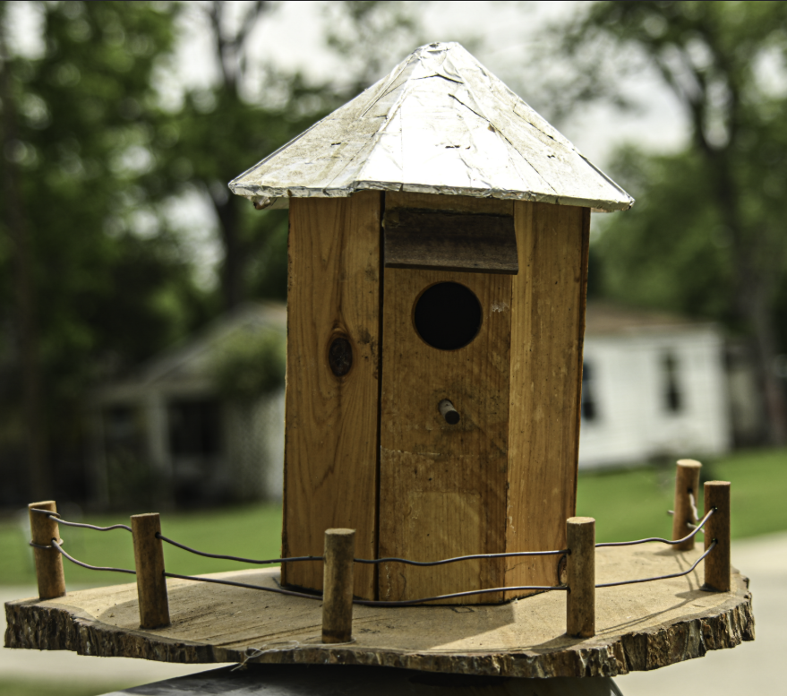 Wooden birdhouse with a metal roof on a small fenced platform outdoors, against a blurred green background