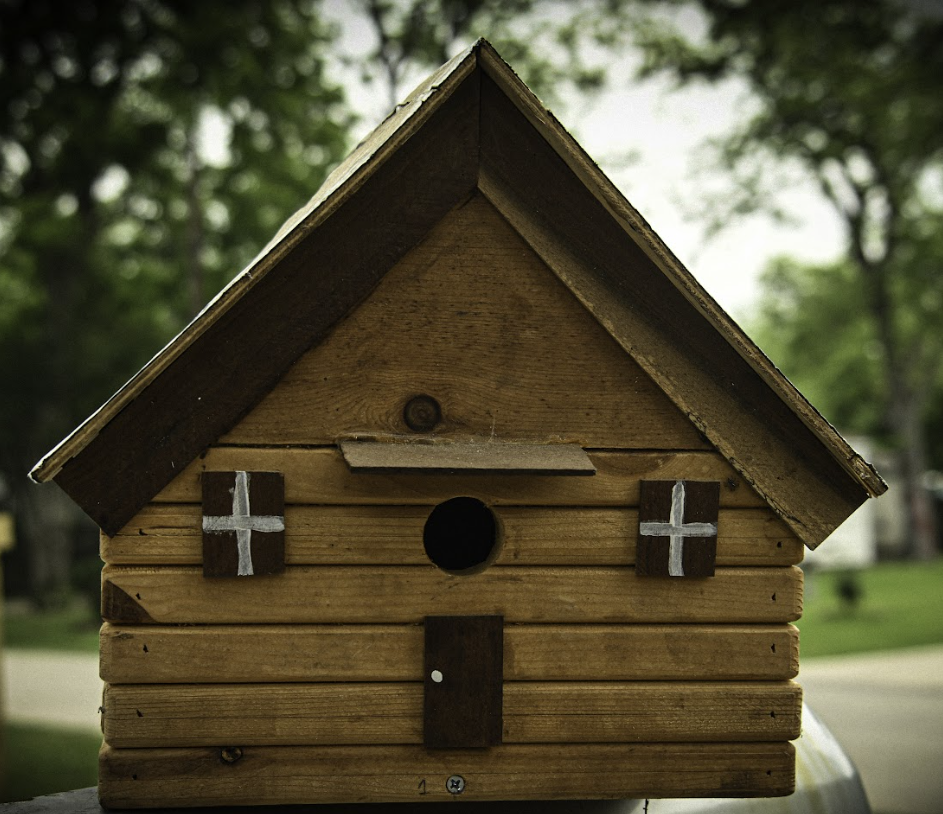 Wooden birdhouse with a steep roof, round opening, and white cross decorations, set outdoors among trees