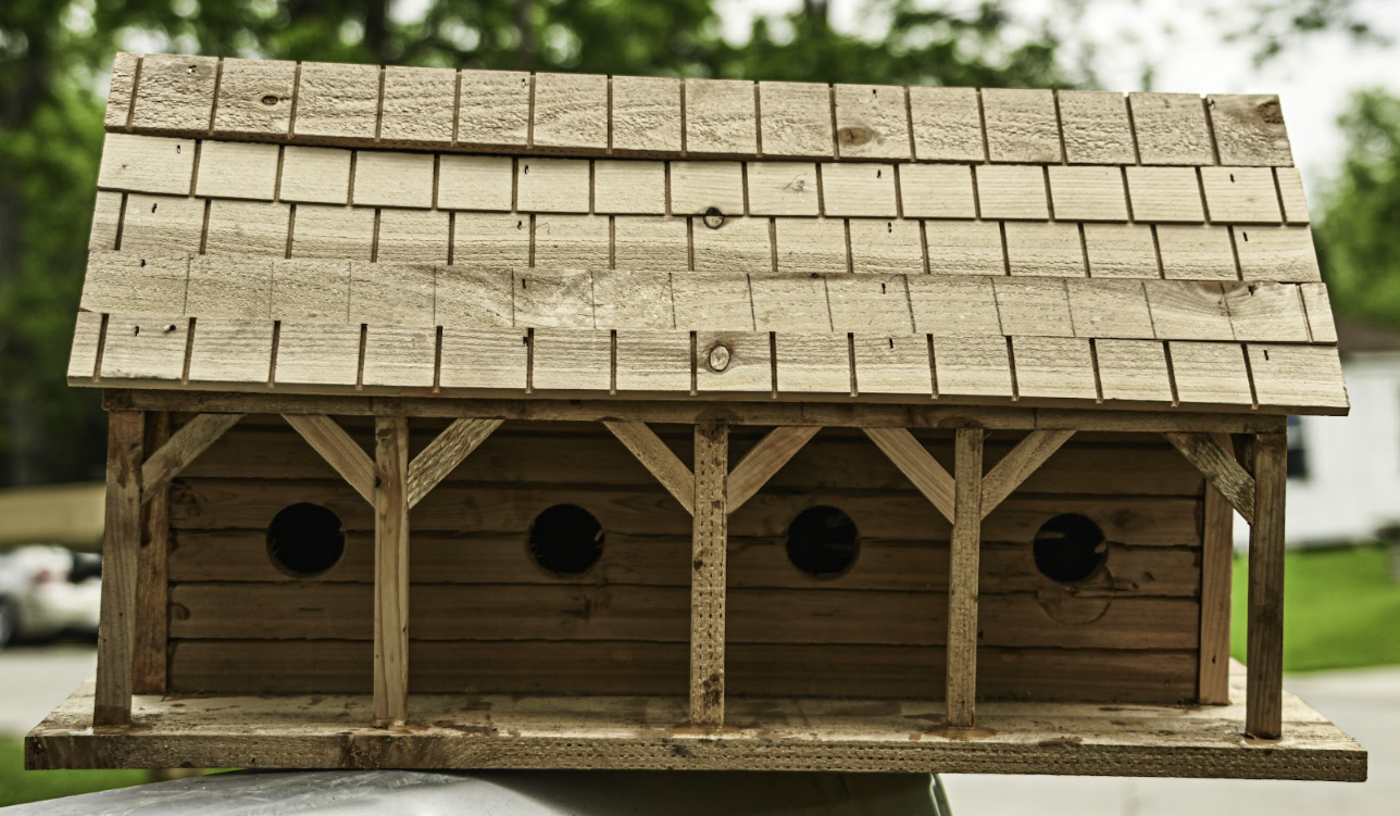 Wooden birdhouse with a shingled roof and four round openings, sitting outdoors on a surface.