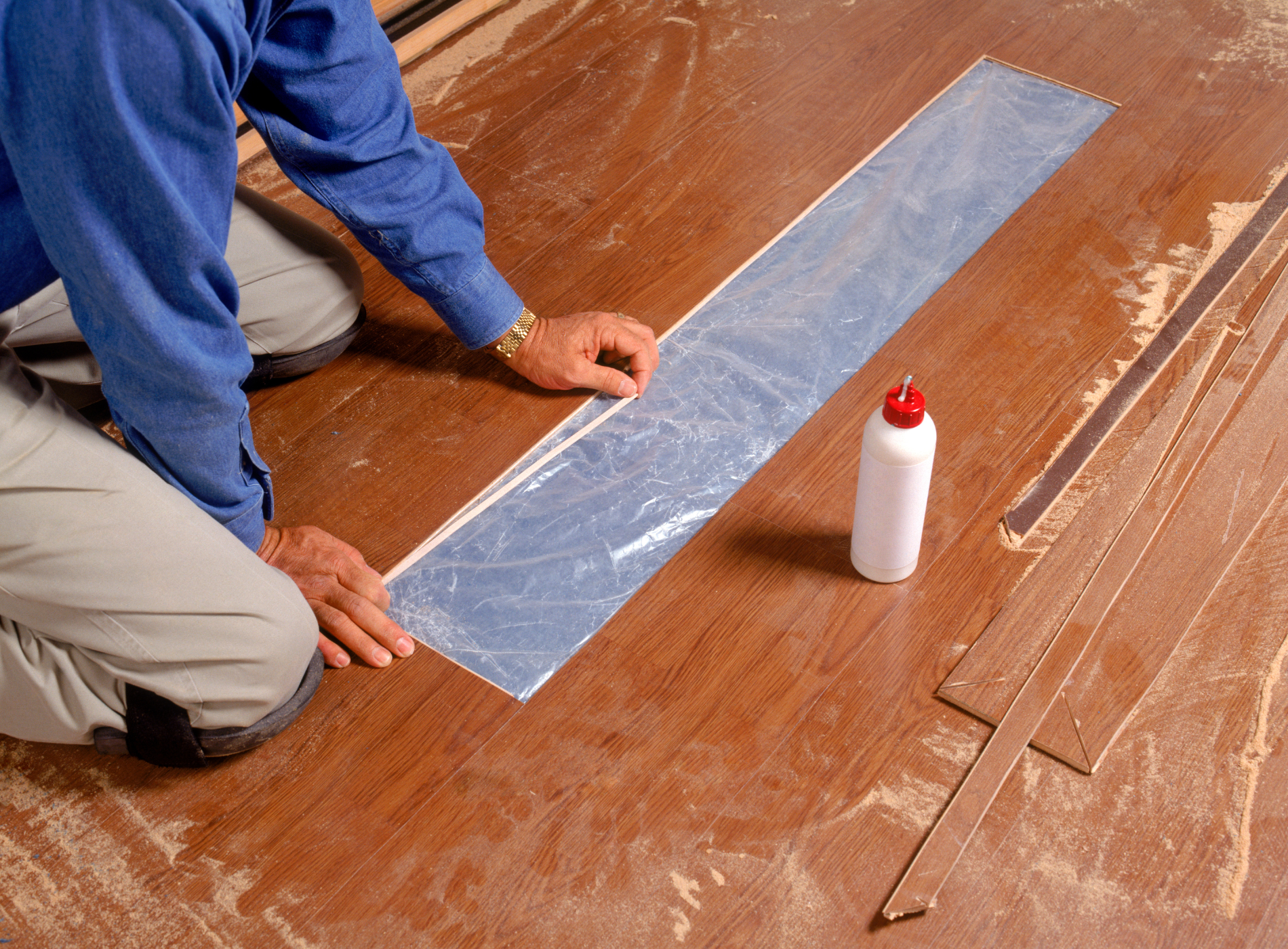 A man is kneeling on a wooden floor next to a bottle of glue