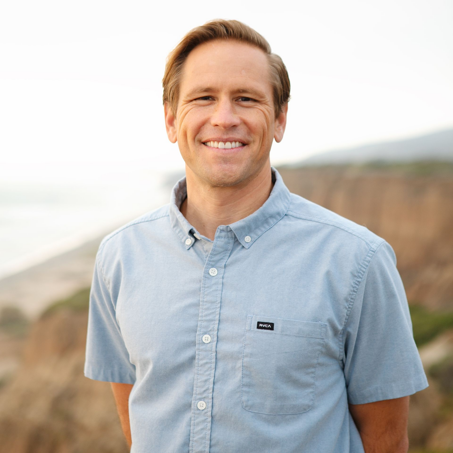 Man with fair skin smiles outdoors in a blue button-down shirt; a beach and cliffside are in the background.