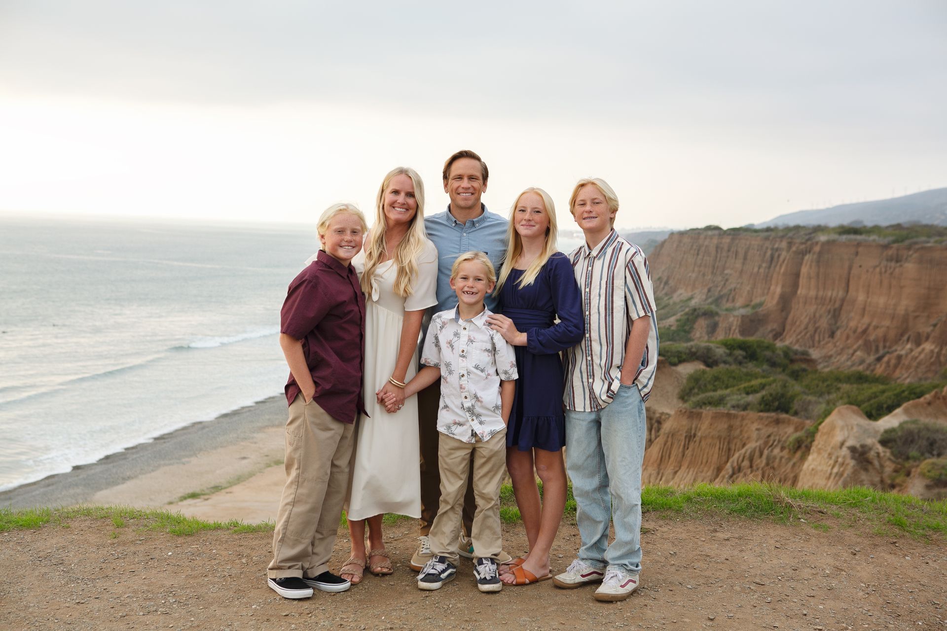 Family of six stands on a cliff overlooking the ocean; cloudy sky; neutral tones.