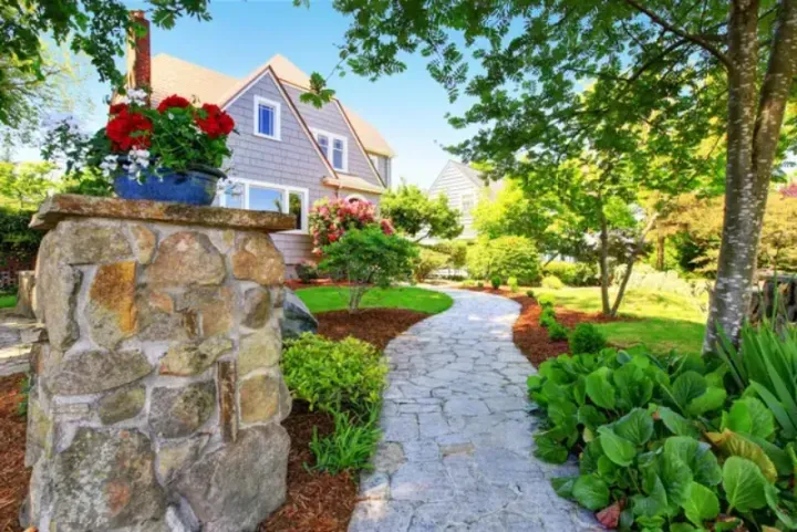 A stone walkway leading to a large house surrounded by trees and bushes.