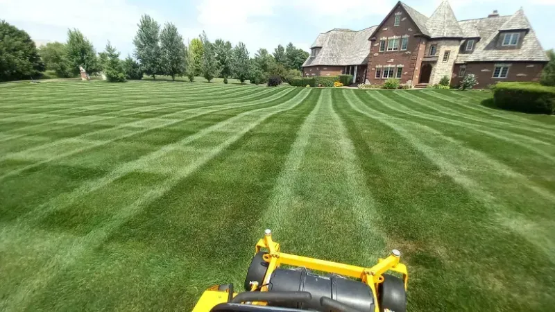 A yellow lawn mower is cutting a lush green lawn in front of a large house.