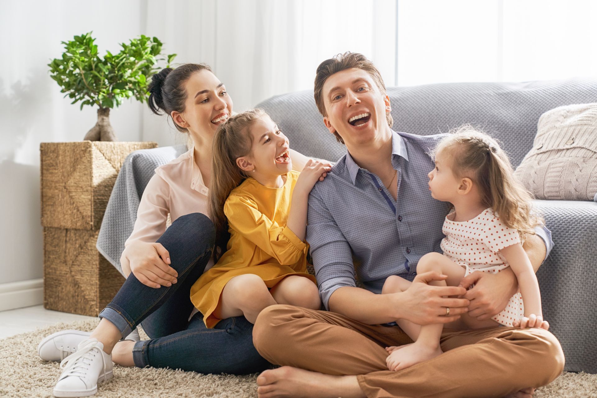 Family laughing together on a living room floor, with a sofa and potted plant in the background