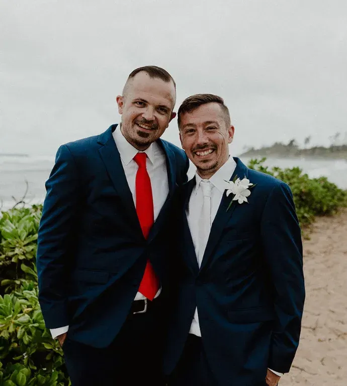 Two smiling men in navy suits, one with a red tie, standing outdoors by the water.