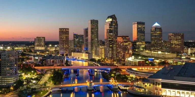 City skyline at dusk with illuminated skyscrapers, bridges, and reflections over a river