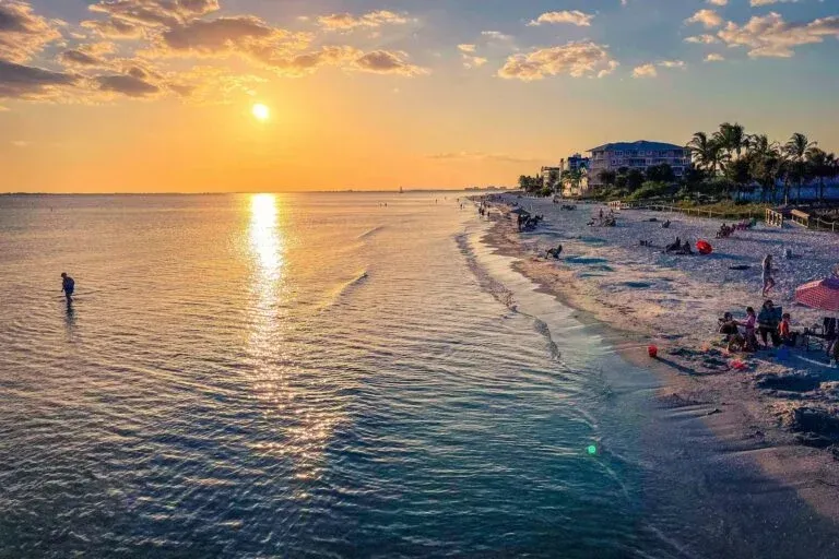 Sunset over calm ocean beside a sandy beach with a few people and palm-lined shoreline