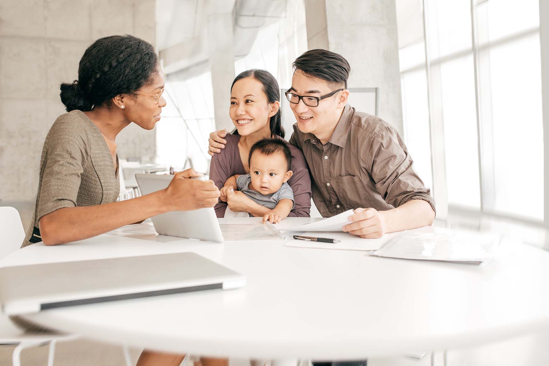 Three people meeting with a baby at a bright office table, smiling and discussing paperwork