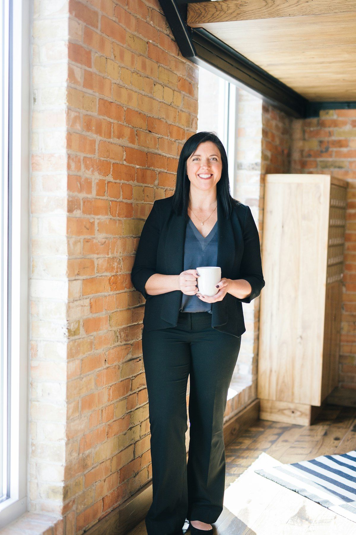 Woman standing in a brick-walled room holding a white mug, beside a large window.
