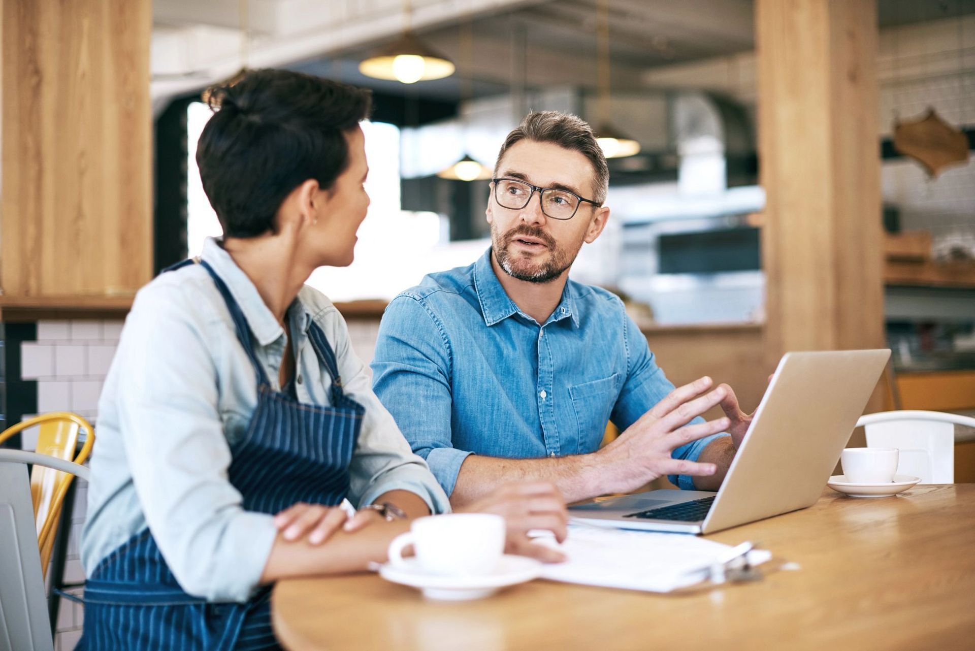 Two coworkers talking at a table with a laptop and coffee in a bright café
