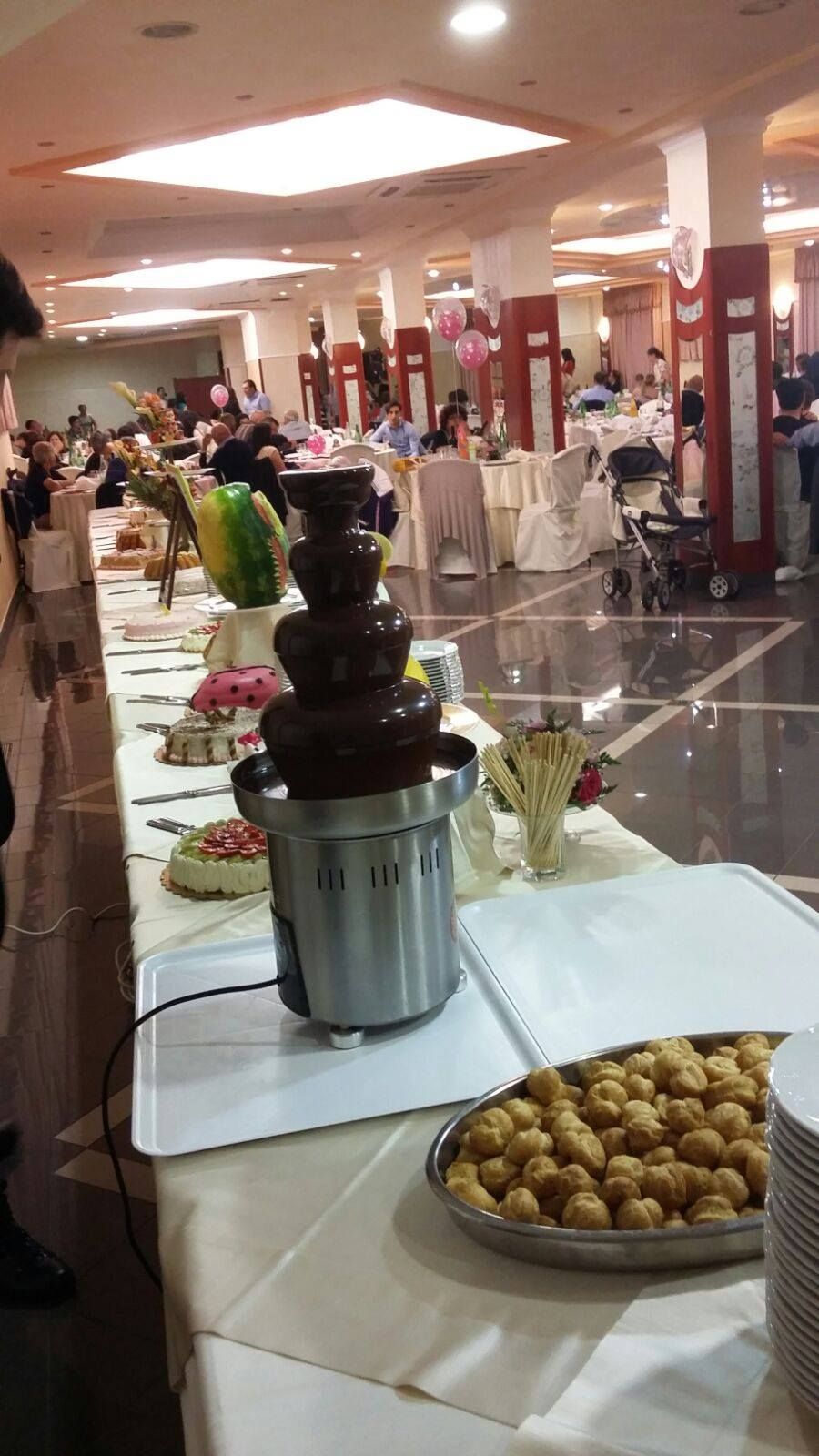 Fontana di cioccolato su un tavolo da buffet durante un evento, con pasticcini e persone sullo sfondo.