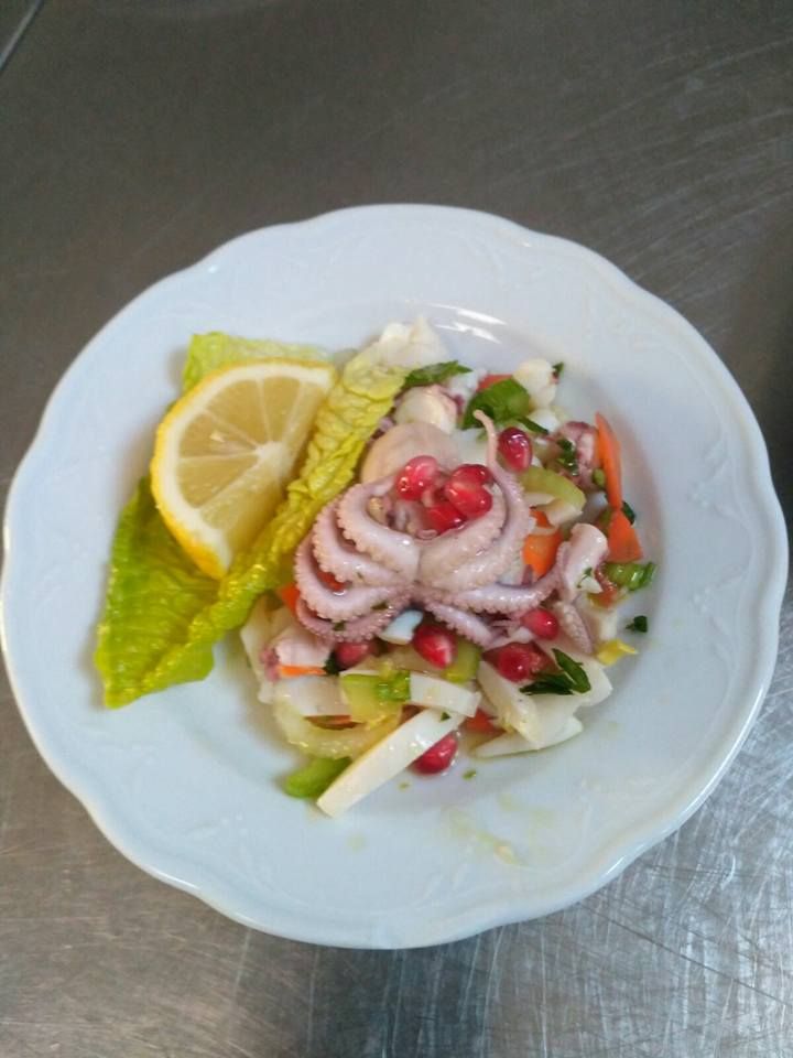 Insalata di polpo con verdure, semi di melograno e spicchio di limone su un piatto bianco.