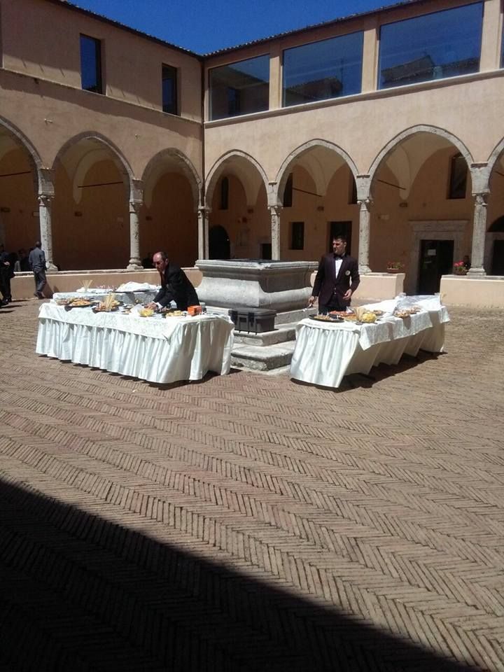 Due persone sono in piedi dietro tavoli pieni di cibo in un cortile con passaggi ad arco.