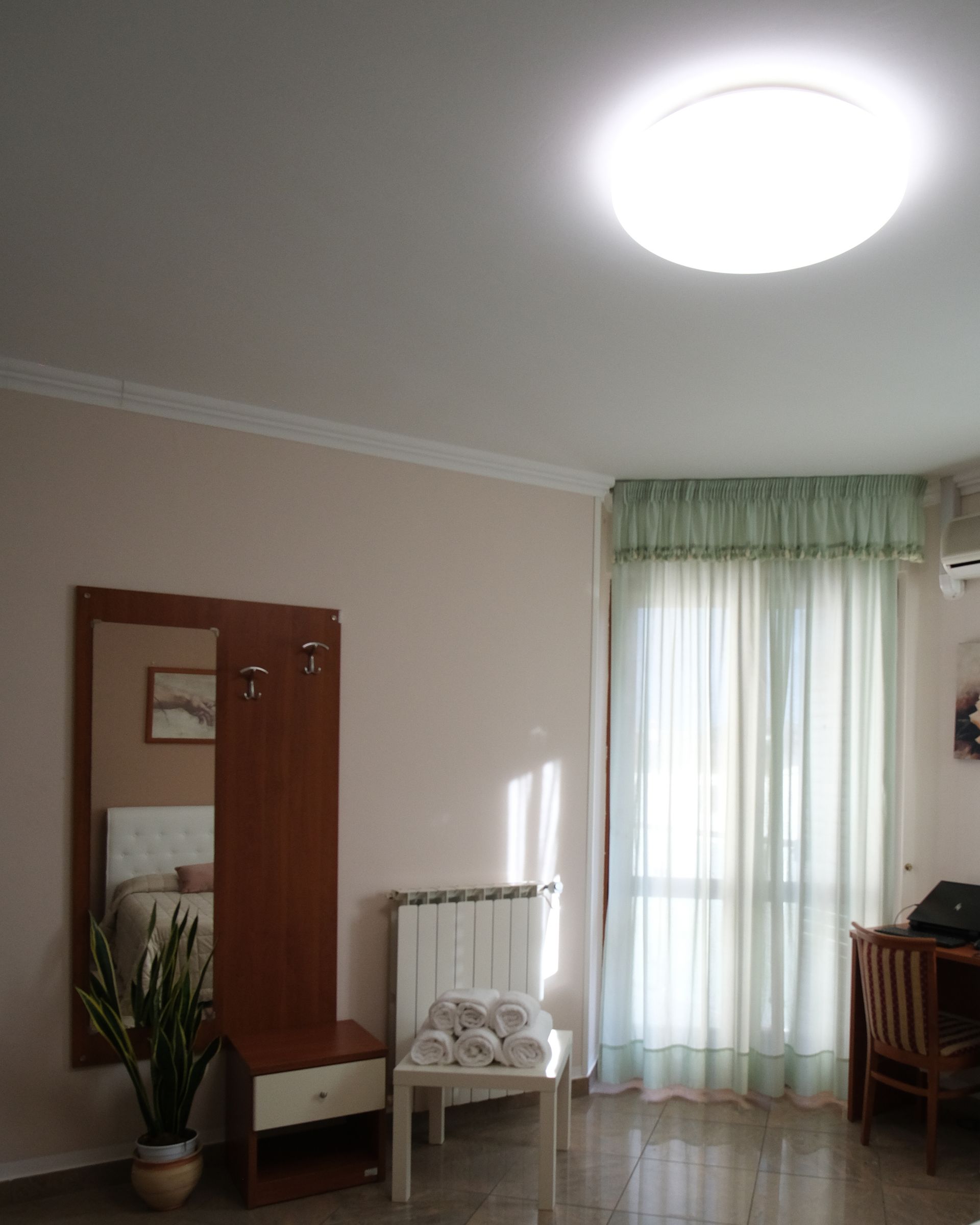 Vista interna di una stanza con specchio, attaccapanni, sedia con asciugamani e finestra con tende.