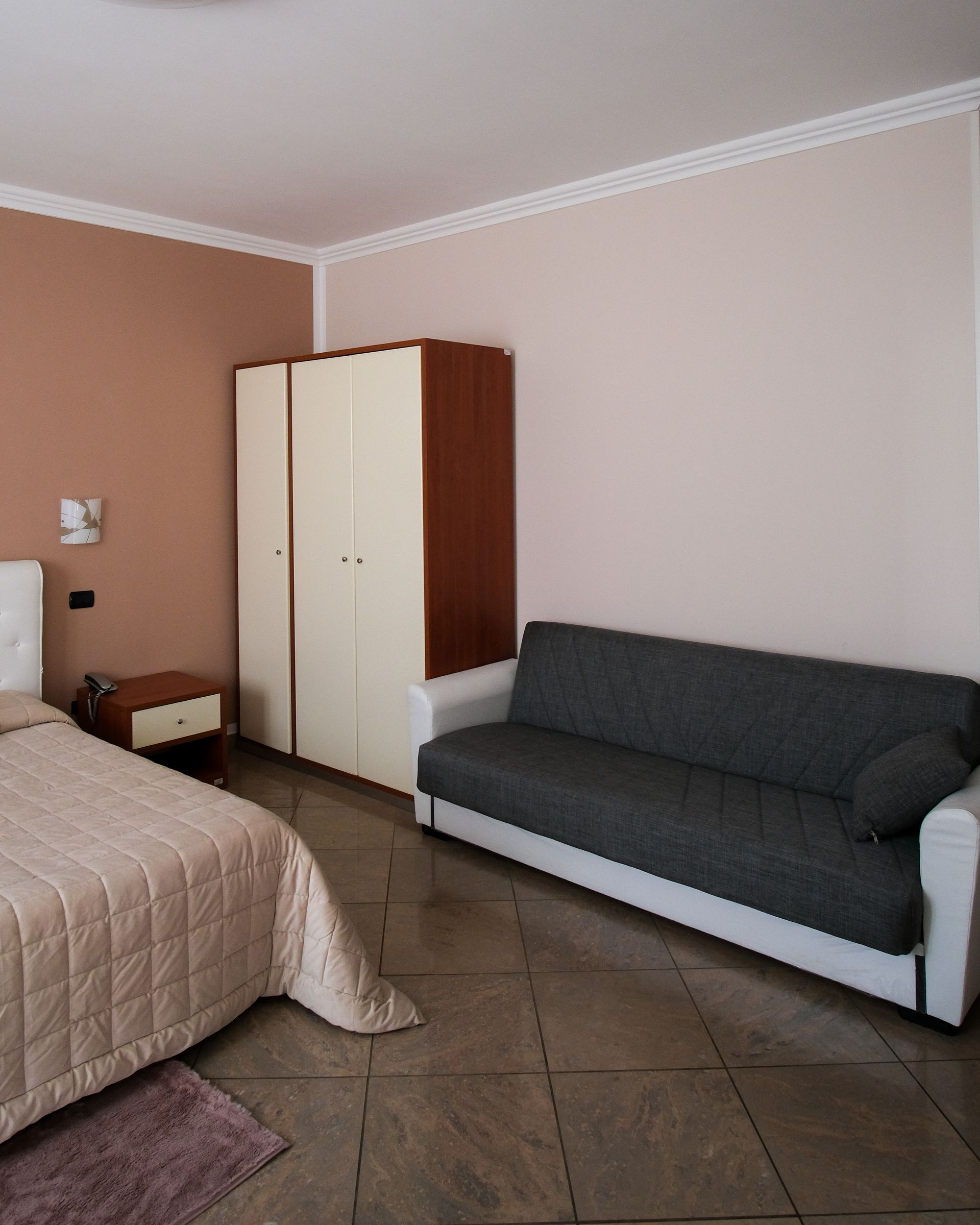Camera da letto con letto, armadio e divano. Colori beige, marrone e bianco. Pavimento piastrellato.