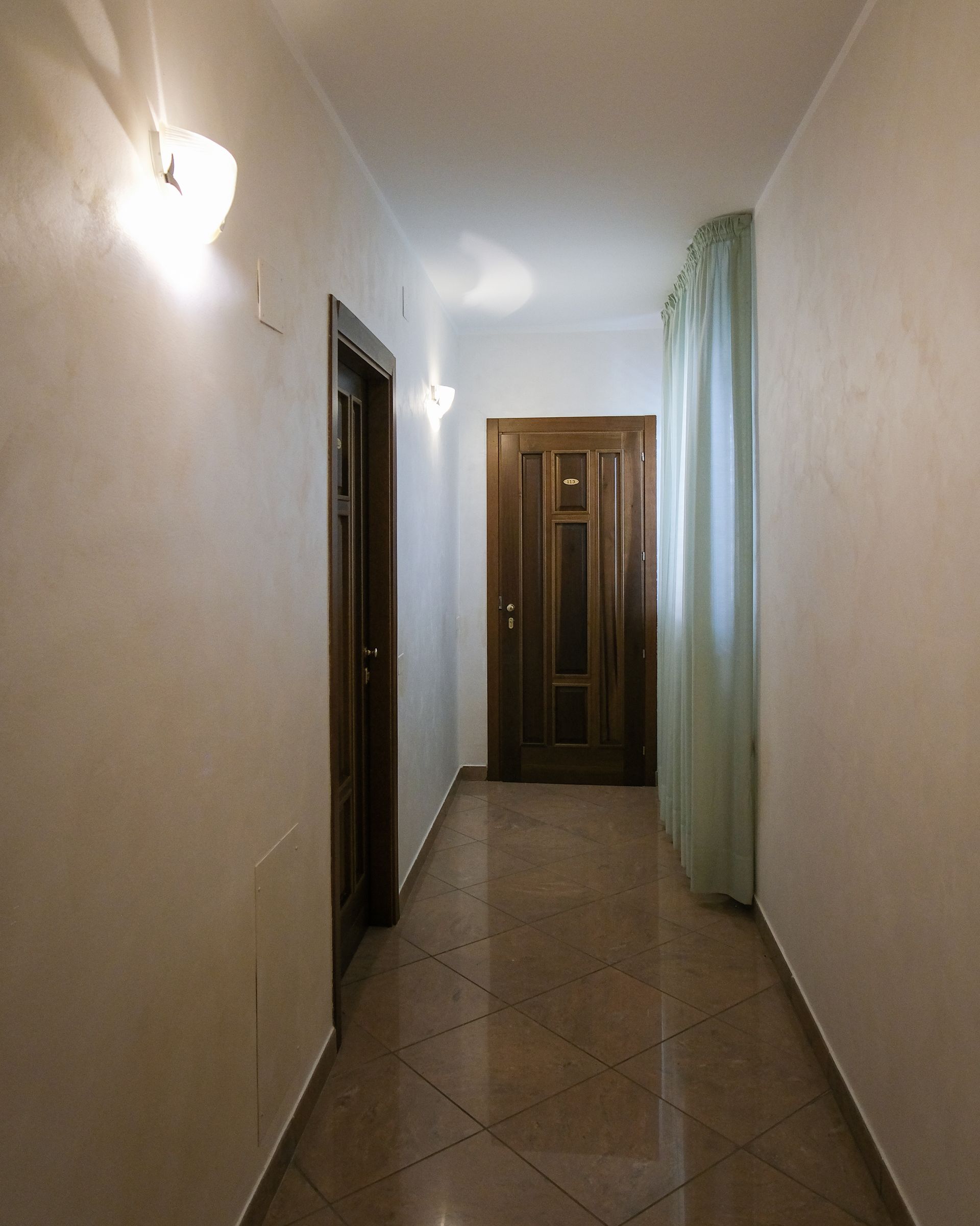 Corridoio stretto con pareti bianche, pavimento in piastrelle marroni e porte in legno.