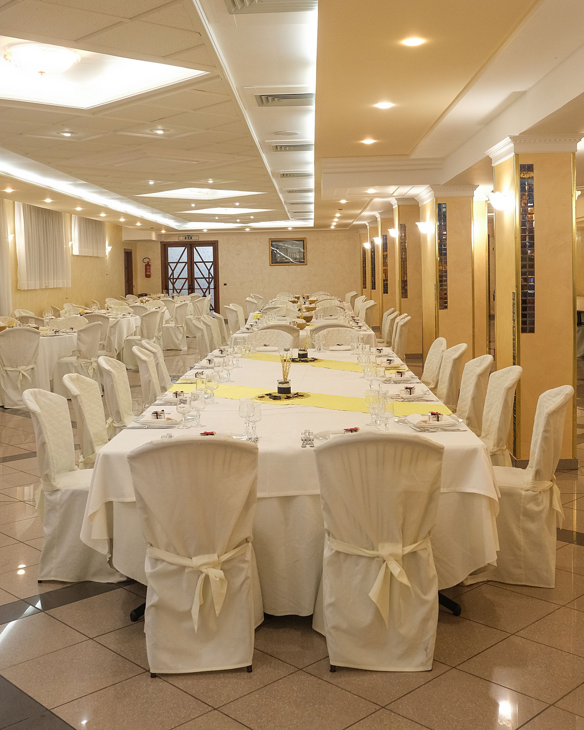 Lungo tavolo rettangolare da banchetto apparecchiato per un evento formale in una stanza piena di luce; sedie e tovaglie bianche.