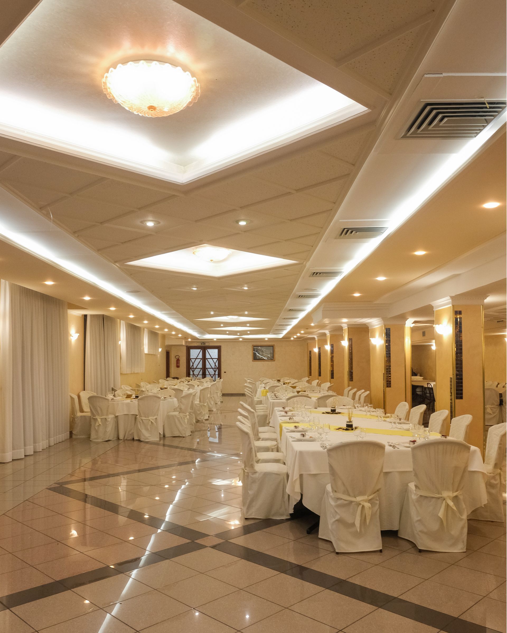 Sala per banchetti con tavoli ricoperti di tovaglie bianche, pronta per un evento, illuminata in modo decorativo.