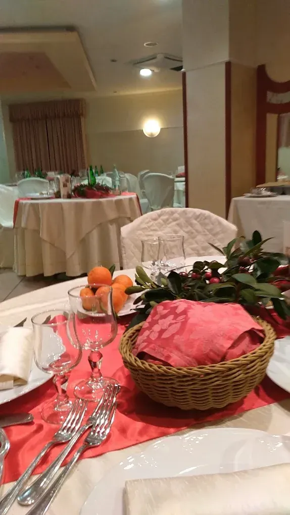 Tavolo del ristorante apparecchiato con runner rosso, cestino, frutta, bicchieri e posate. Altri tavoli sullo sfondo.