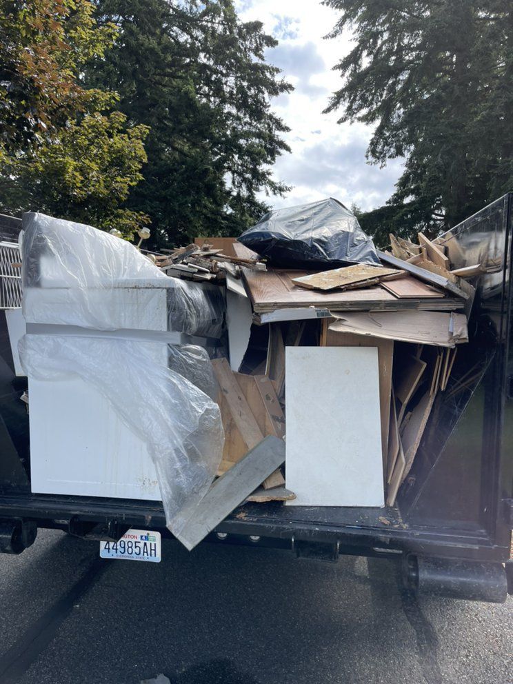 A dumpster filled with construction debris: wood, tiles, and covered items, against a tree background.