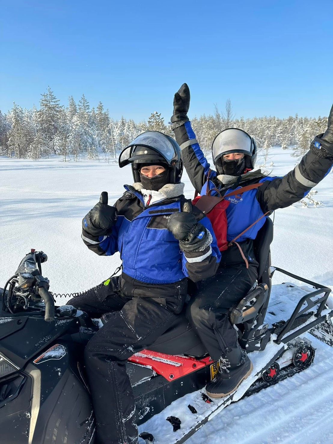 Two people on a snowmobile in a snowy forest, both wearing helmets and winter gear, raising hands with thumbs up.