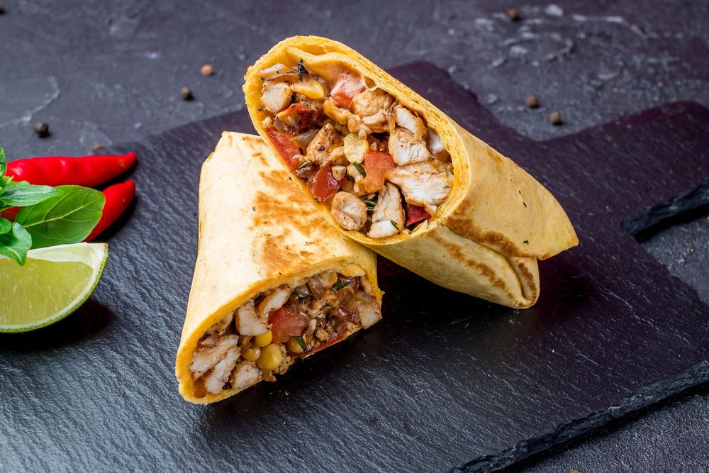 Two grilled wrap halves filled with diced meat and vegetables on a dark slate board, with lime and chili peppers. Two grilled wrap halves filled with diced meat and vegetables on a dark slate board, with lime and chili peppers.