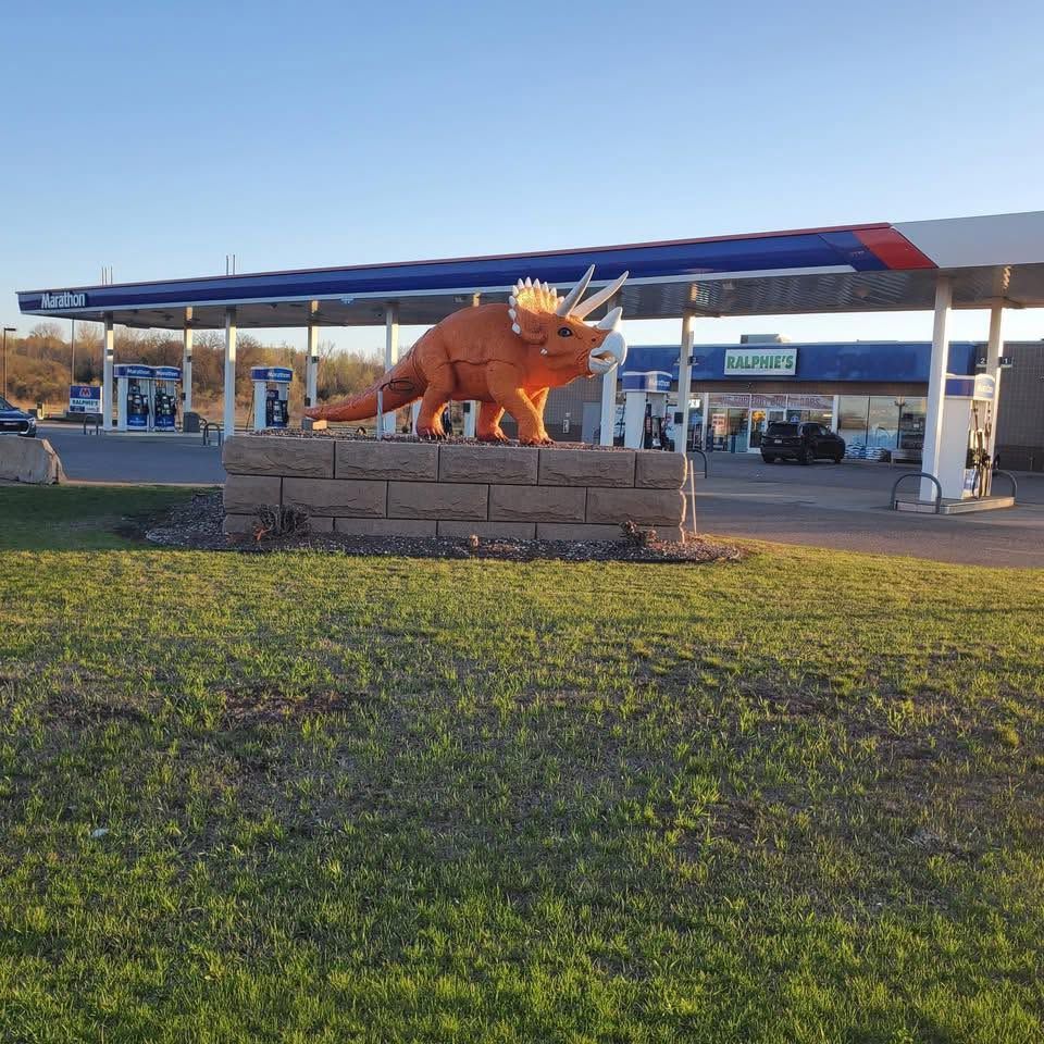 Orange triceratops sculpture on rocks beside a grassy field under a blue sky