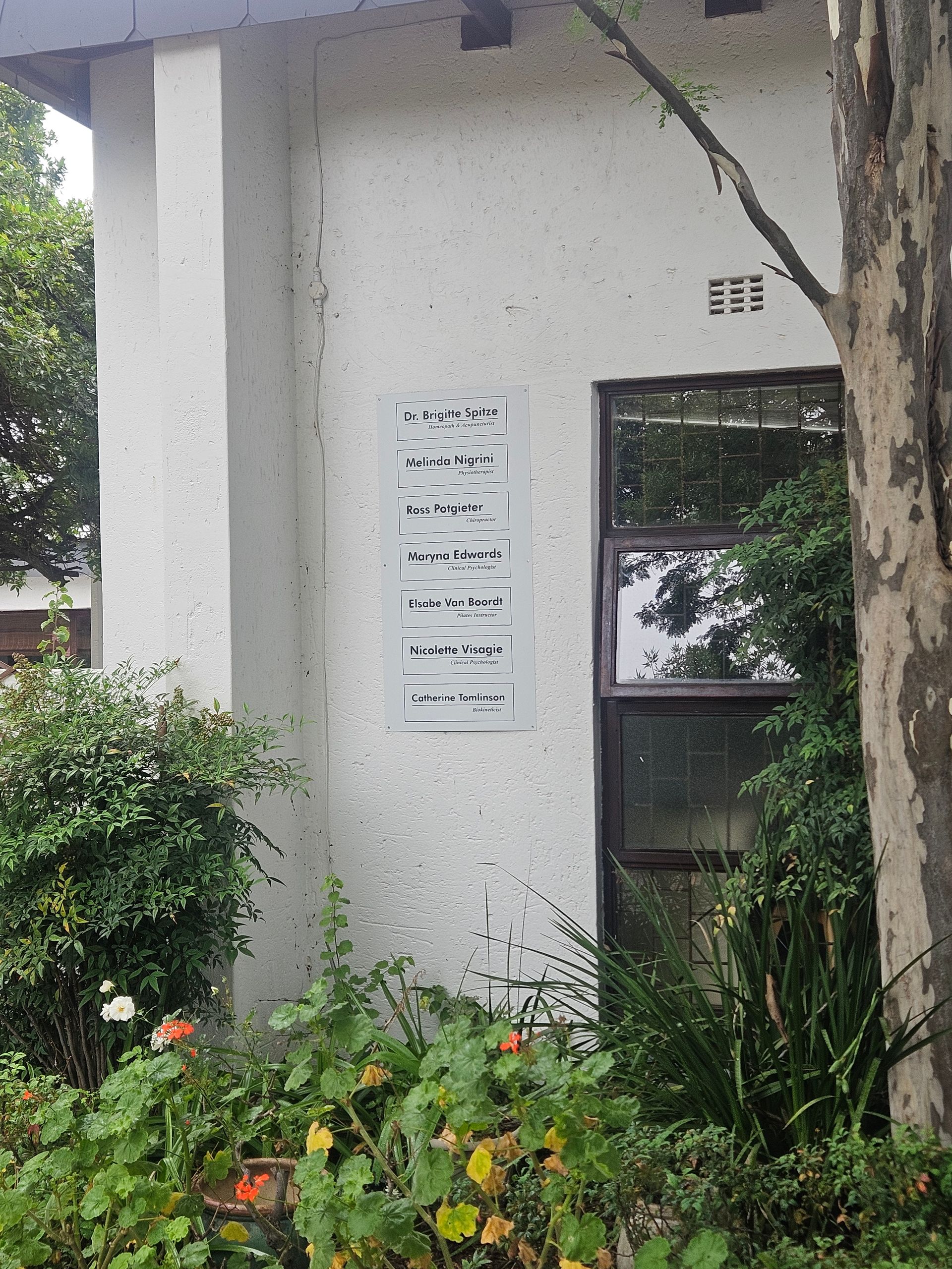 A white exterior wall of a building features a vertical directory sign next to a window, surrounded by green shrubs.
