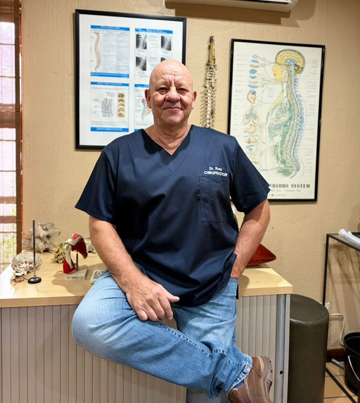 A professional in dark blue scrubs sits on a desk in a clinic, with anatomical posters on the wall behind them.