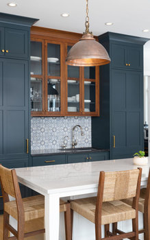 Modern kitchen with blue cabinets, white island, glass-front hutch, and two woven chairs under pendant light
