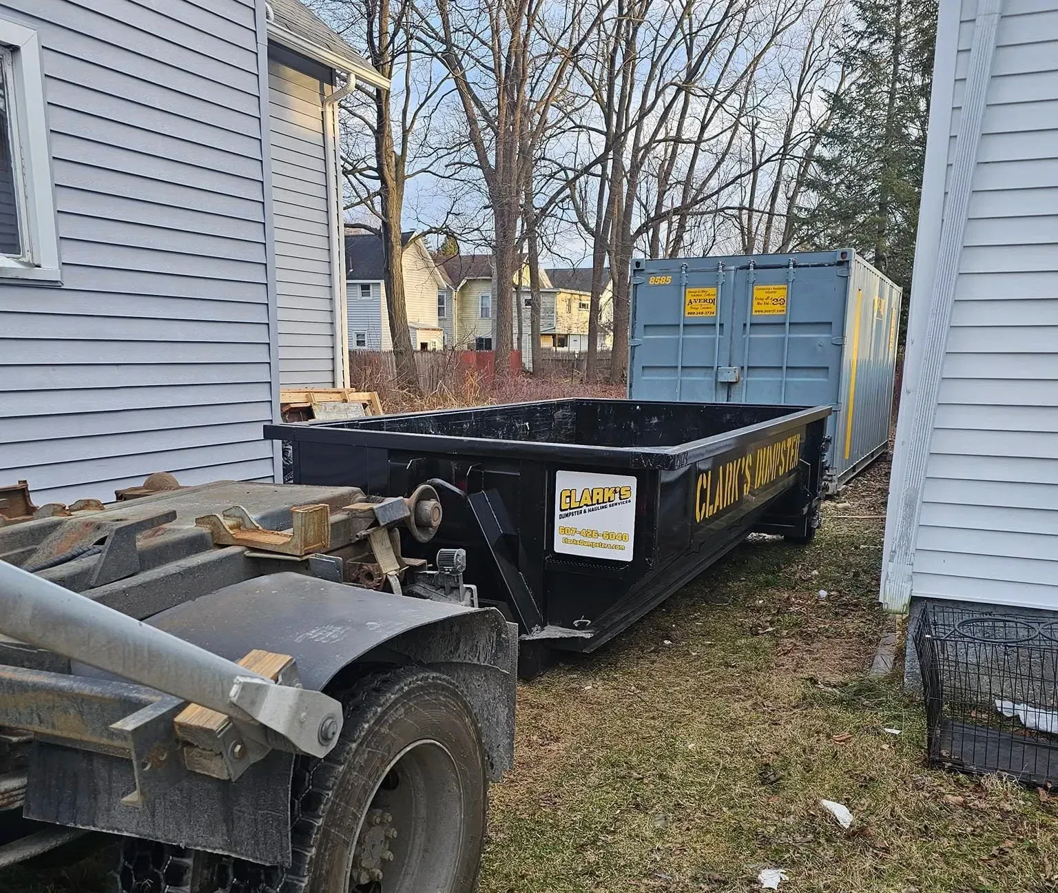 A dumpster is sitting next to a truck in front of a house.