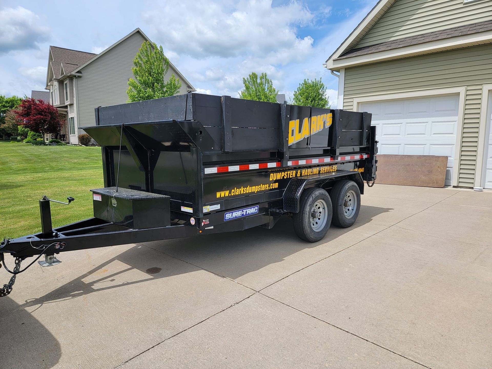 A black dump trailer parked on a concrete driveway in front of a house on a sunny day.
