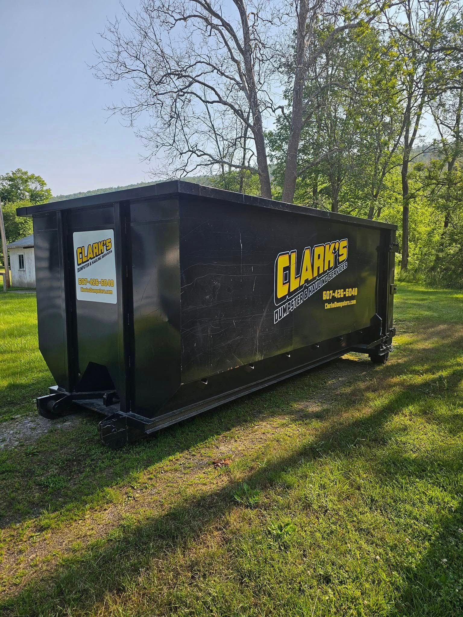 A large black Clark’s dumpster sitting on grass in a wooded area.