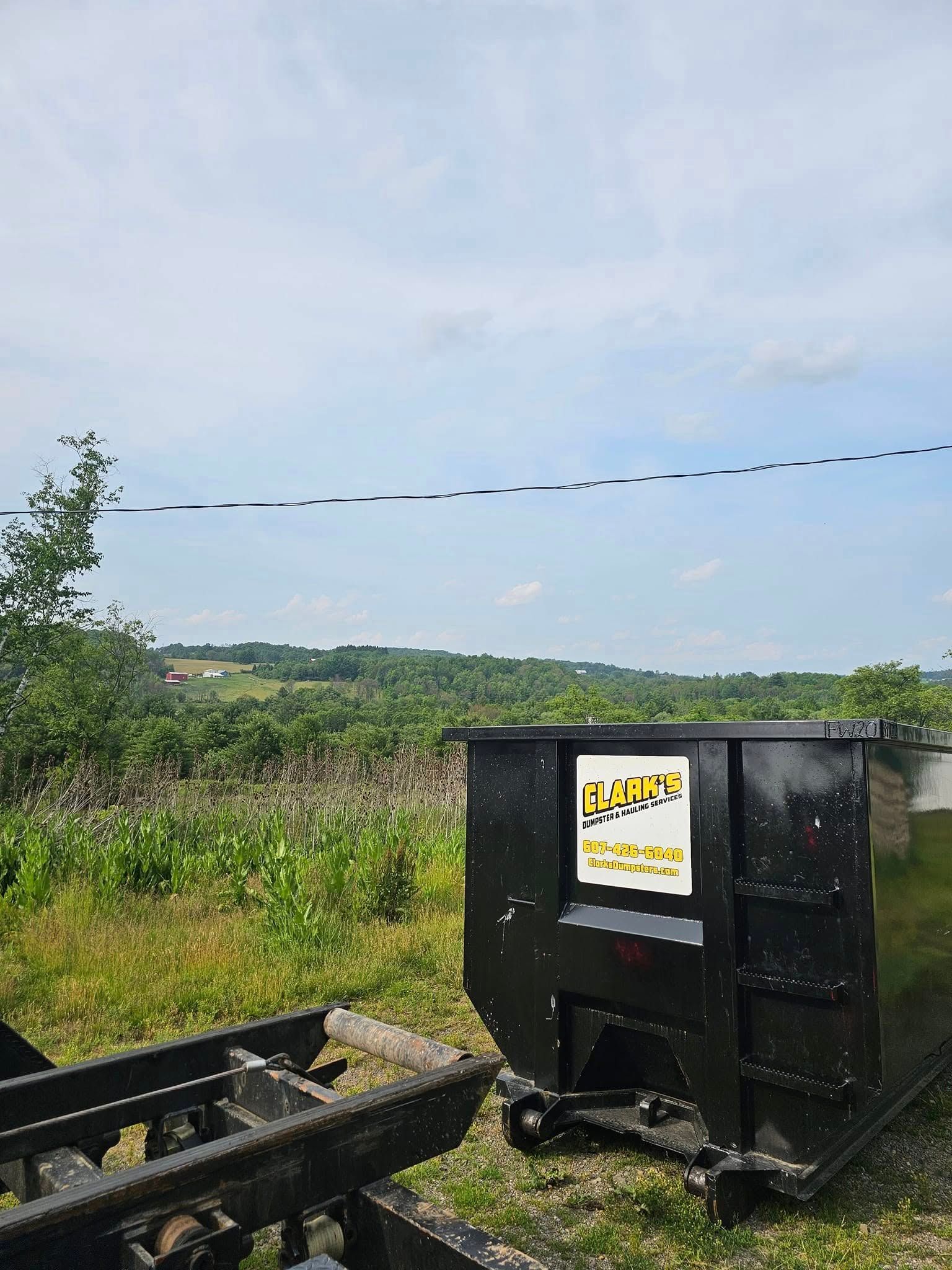 A black waste dumpster stands in a grassy, open field overlooking a rolling, tree-covered landscape under a cloudy sky.