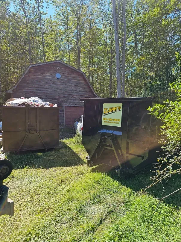 Two dumpsters sit on grass in front of an old, weathered wooden barn surrounded by trees on a sunny day.