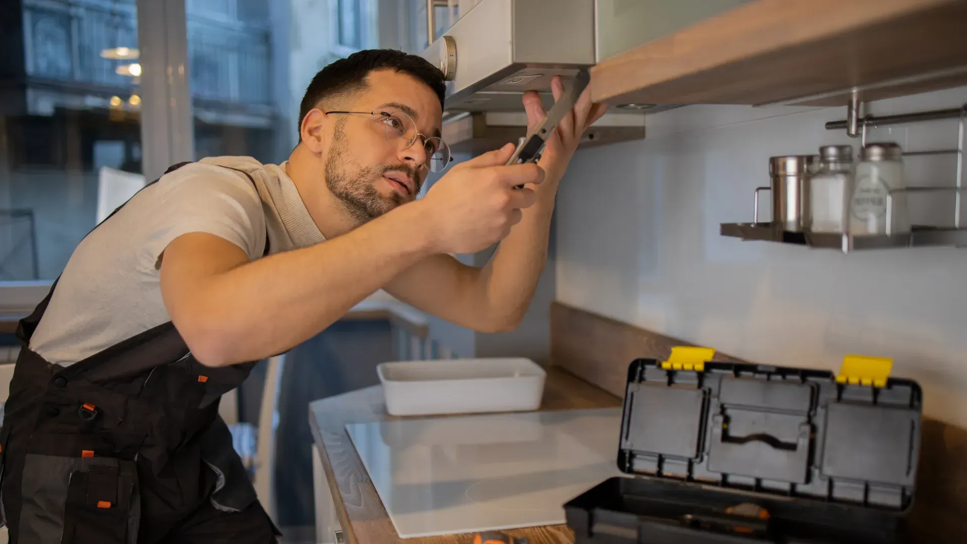 A man in overalls fixes a kitchen appliance with a screwdriver, toolbox nearby.